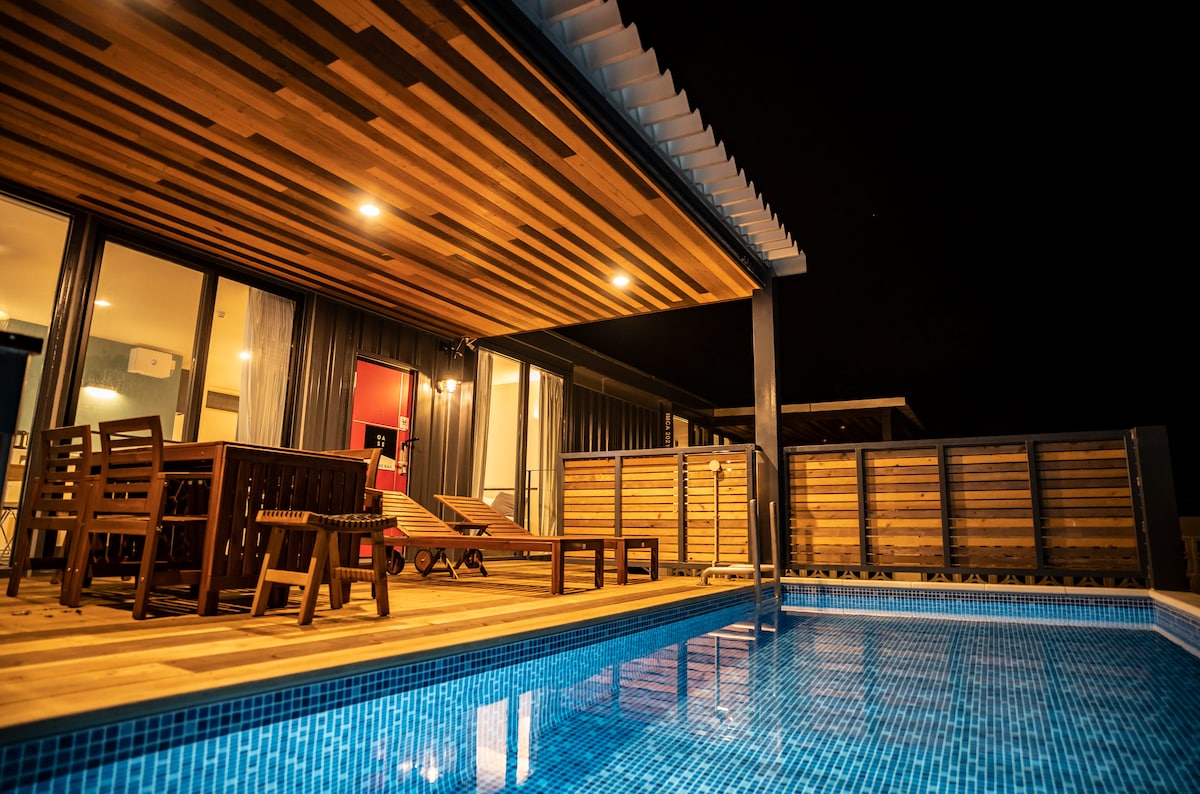 A serene outdoor space features a shimmering pool illuminated by soft lighting. A wooden terrace is equipped with lounging chairs and a dining area, creating an inviting setting for relaxation and social gatherings. The modern structure is highlighted against the night sky.