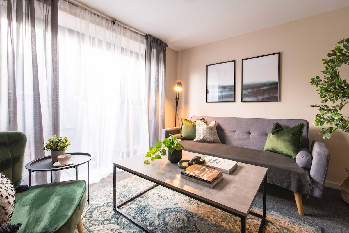 A bright living area is showcased, featuring a comfortable grey sofa accented by green pillows. A coffee table is positioned centrally, adorned with a plant and decor items. Sheer curtains frame a large window, allowing natural light to fill the space. Two green chairs enhance the cozy atmosphere.