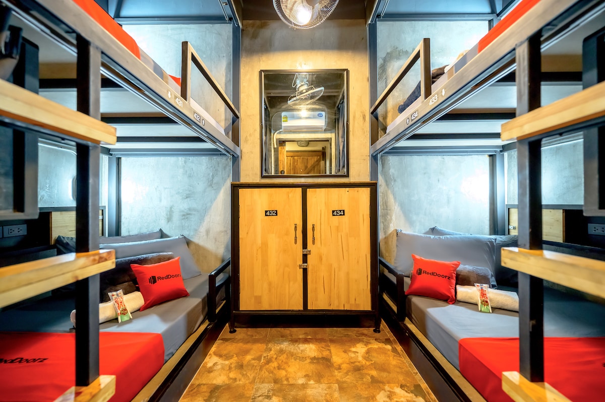 A modern shared sleeping area features two bunk beds arranged against the walls, with plush gray bedding and vibrant red accent pillows. A mirrored area offers additional lighting, and wooden cabinets provide storage solutions. The industrial design is complemented by warm, neutral tones.