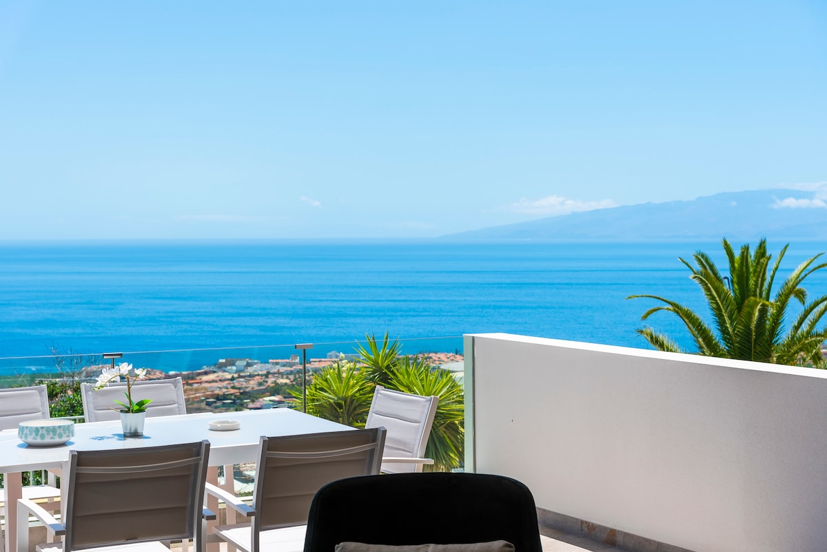 An outdoor terrace showcases a dining table with six chairs, overlooking the expansive Atlantic Ocean. Lush greenery is visible in the foreground, while the distant island of La Gomera is framed by a clear blue sky.