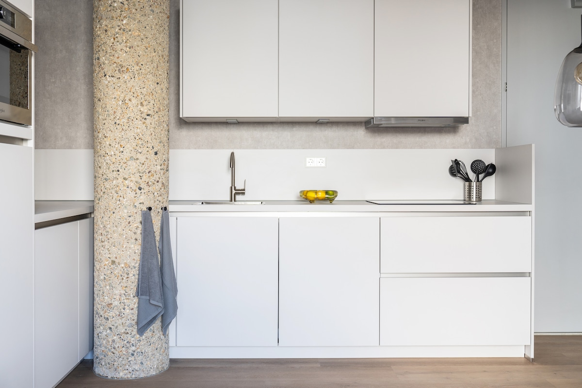A modern kitchen area features sleek white cabinetry and a textured column. Stainless steel appliances are present, including an oven and a sleek faucet. A bowl of fruits adds a touch of color, while neatly arranged kitchen utensils are visible, creating a functional cooking space.