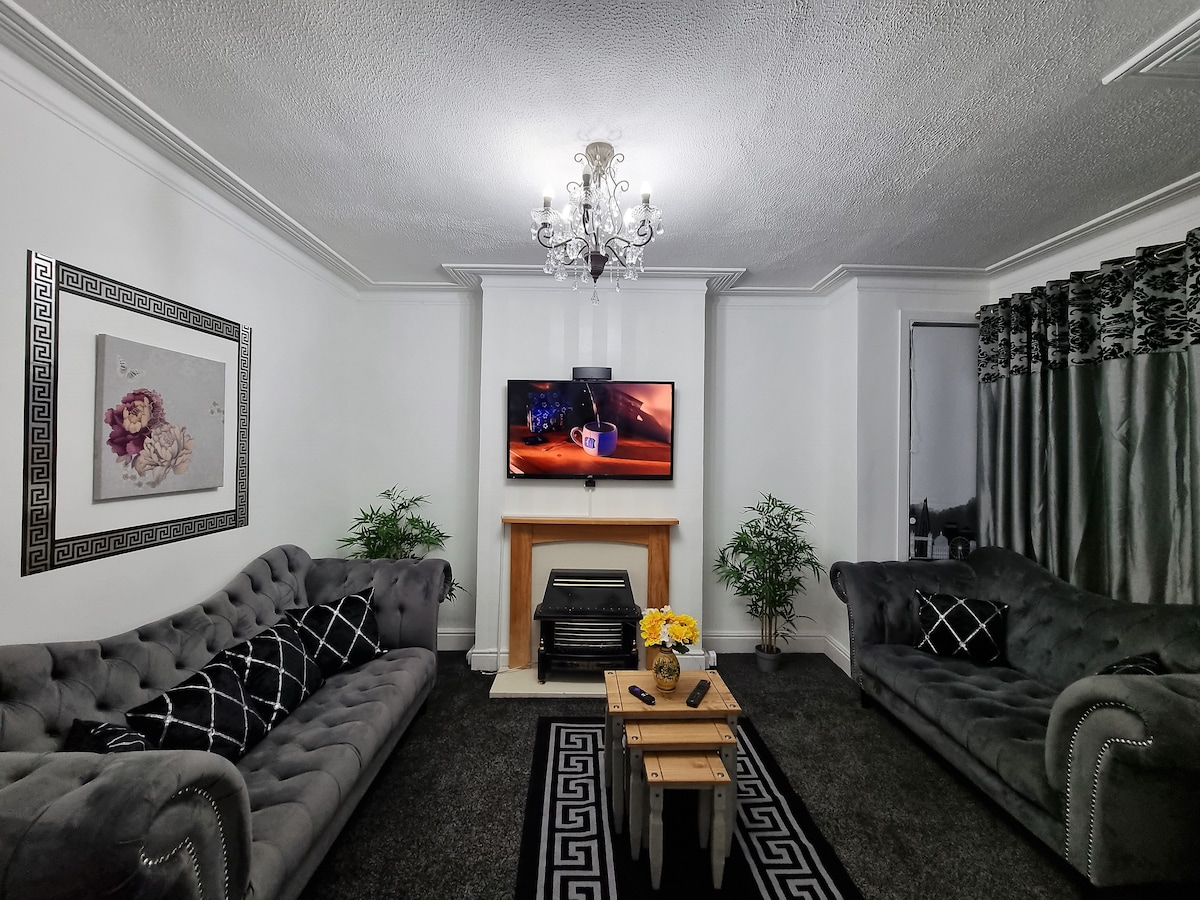 A comfortable living room is presented, featuring two tufted grey sofas facing a central coffee table. A large television is mounted on the wall above a wooden fireplace. Decorative plants are placed in the corners, and curtains frame the windows.