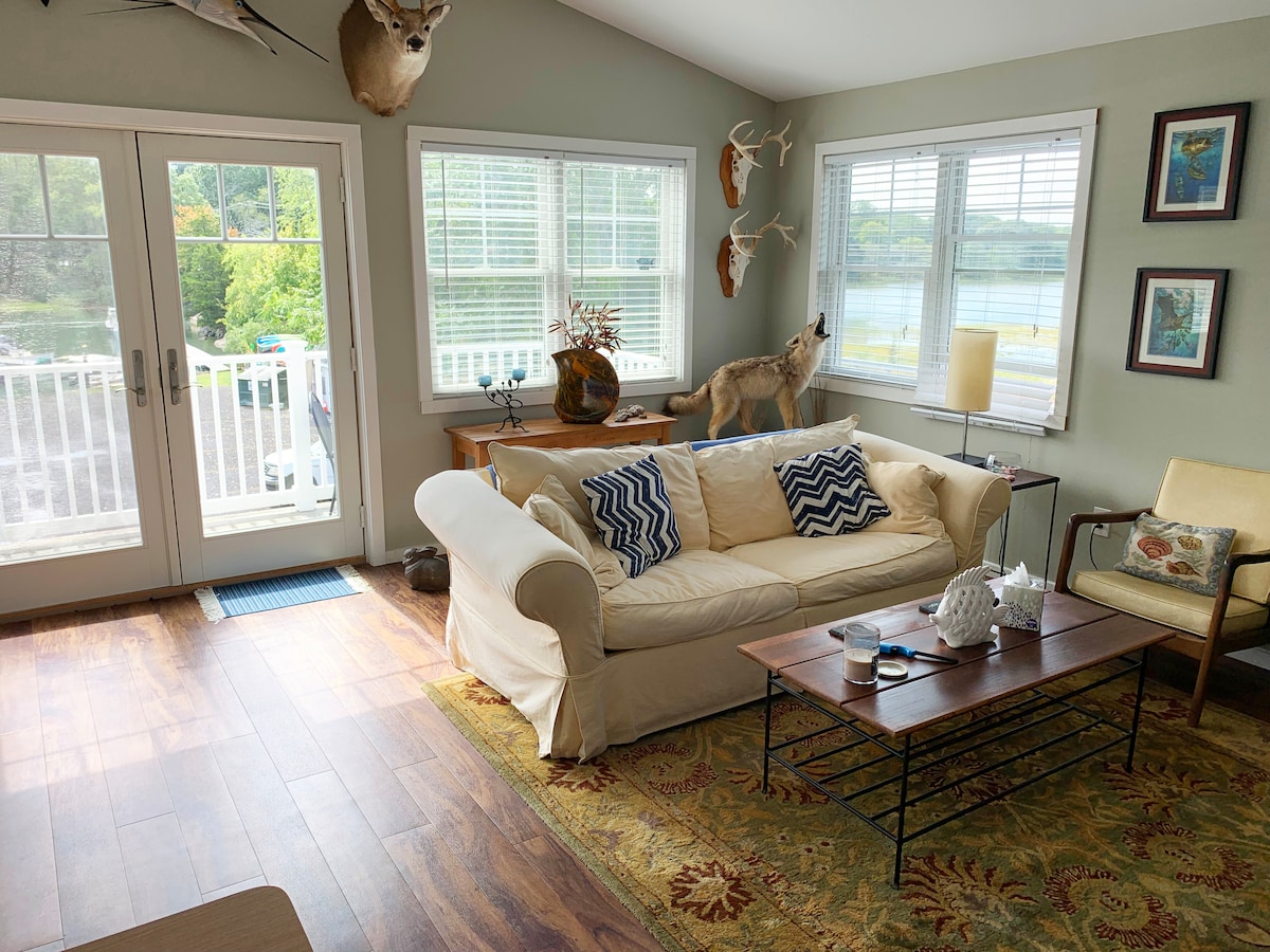 A cozy living area features a cream-colored sofa adorned with patterned pillows, accompanied by a light yellow armchair. Natural light enters through large windows overlooking the water. A wooden coffee table displays decor items, and the space is complemented by a patterned area rug.