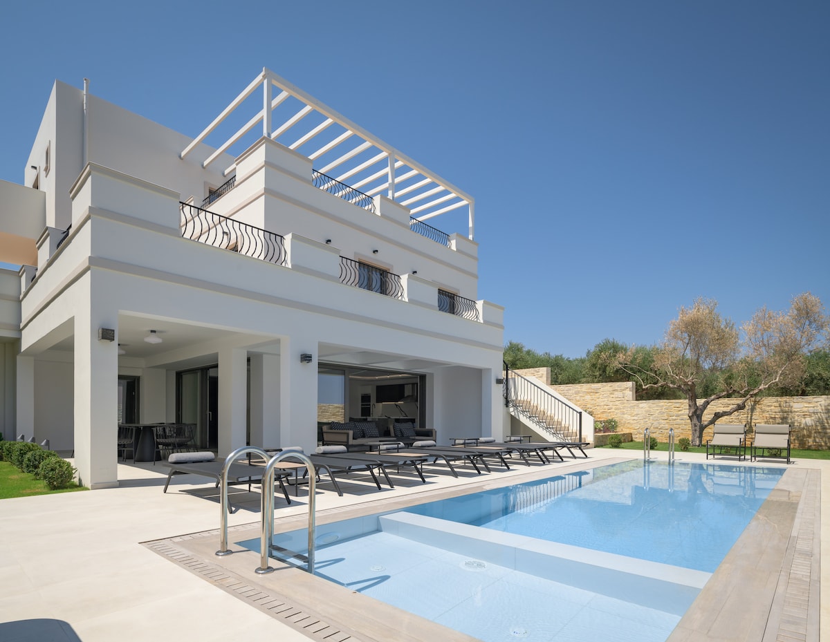 The exterior of the villa showcases a modern design, featuring a spacious terrace with sun loungers and a private swimming pool. The pool area is surrounded by well-maintained gardens, while large sliding doors offer views into the inviting living space.