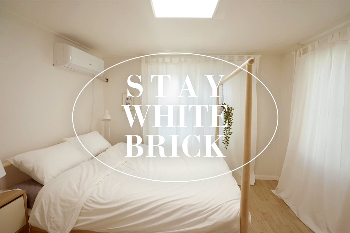 A bright bedroom is highlighted by natural light filtering through sheer white curtains. A spacious bed is dressed in white linens, offering a clean and calming look. An air conditioning unit is mounted on the wall, enhancing comfort during stays.