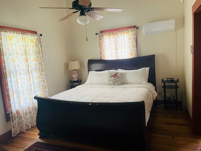 A spacious bedroom features a dark wooden sleigh bed topped with white bedding. Two floral-patterned curtains frame the windows, allowing soft light to enter. A ceiling fan hangs overhead, and a bedside lamp sits on a small table beside the bed.