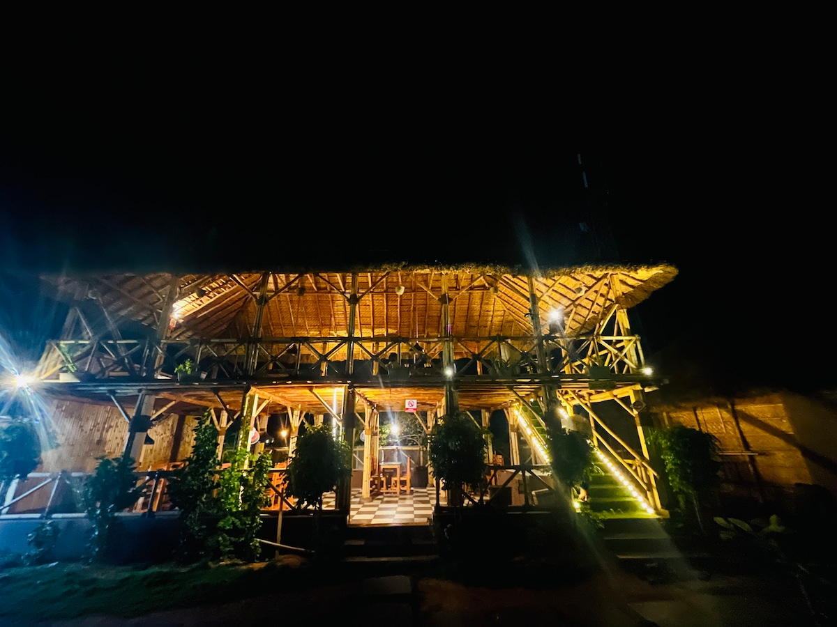 A bamboo cottage is illuminated at night, highlighting its elegant design. The structure features a thatched roof and multiple levels, with outdoor seating areas surrounded by well-maintained greenery. Soft lighting accentuates the inviting atmosphere, creating a warm and welcoming appearance.
