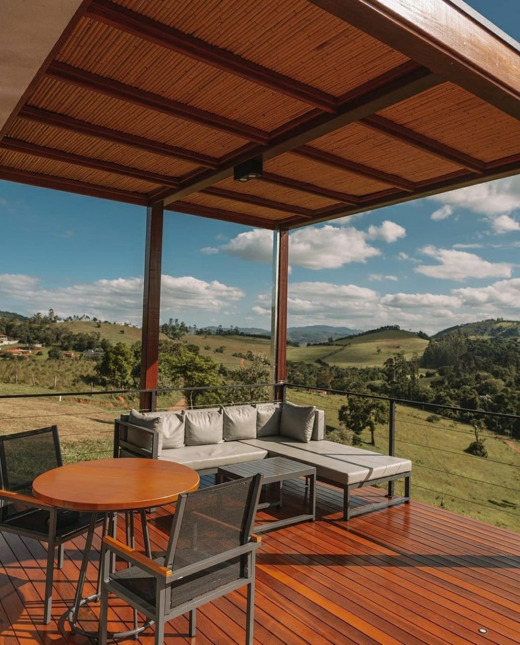 An outdoor seating area is showcased under a covered roof, featuring a circular table and four chairs. A sectional sofa with soft cushions is arranged nearby, overlooking rolling green hills and a clear blue sky. The wooden deck enhances the inviting layout.