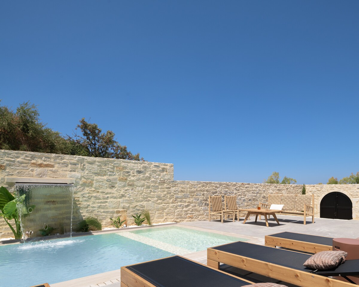 An outdoor area is highlighted by a private swimming pool with a waterfall feature, surrounded by natural stone walls. Comfortable sun beds are arranged nearby, and a relaxing seating area is visible. Clear blue skies above enhance the serene setting.