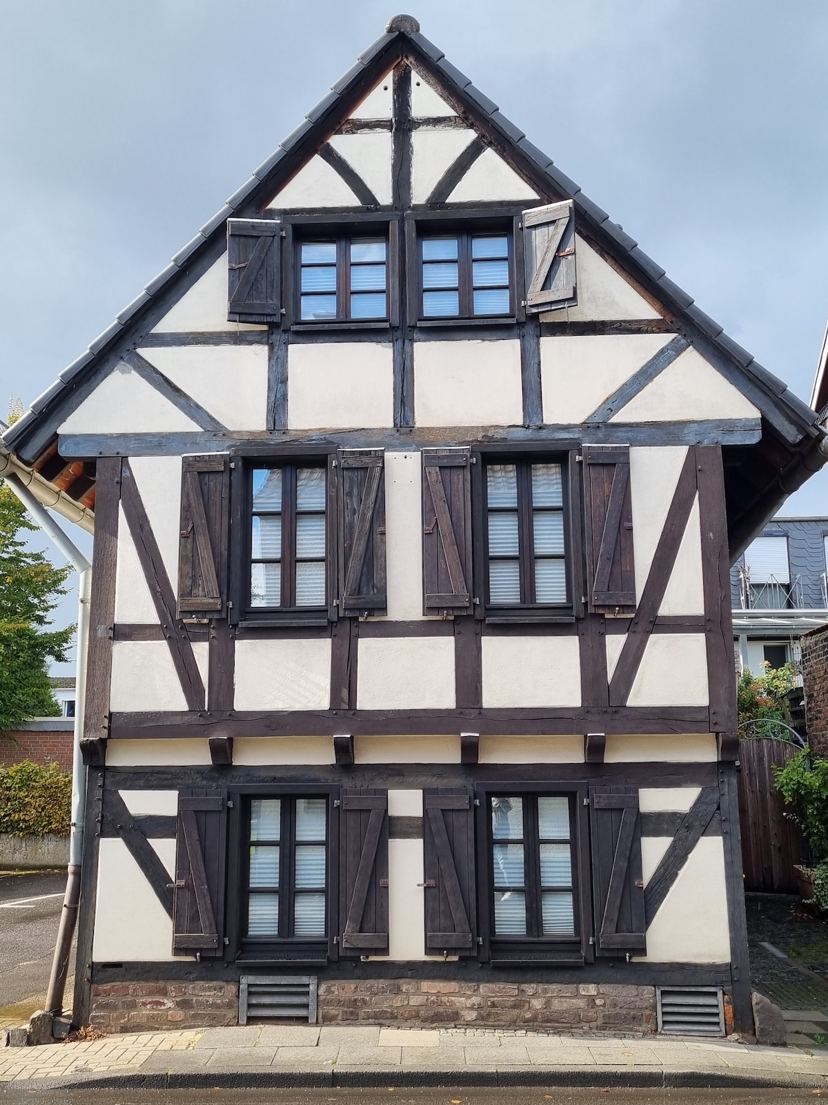The house showcases traditional half-timbered architecture with a white plaster exterior contrasted by dark wooden beams. The facade is characterized by multiple windows, each framed with shutters. The gabled roof adds to its historic charm.