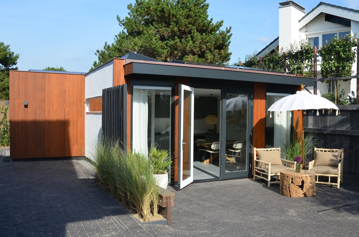 A modern bungalow features large glass doors that open to a private exterior space. Natural wood accents are visible on the façade, complemented by surrounding greenery. Comfortable seating areas are provided on a paved terrace, with an umbrella for shade enhancing the outdoor ambiance.