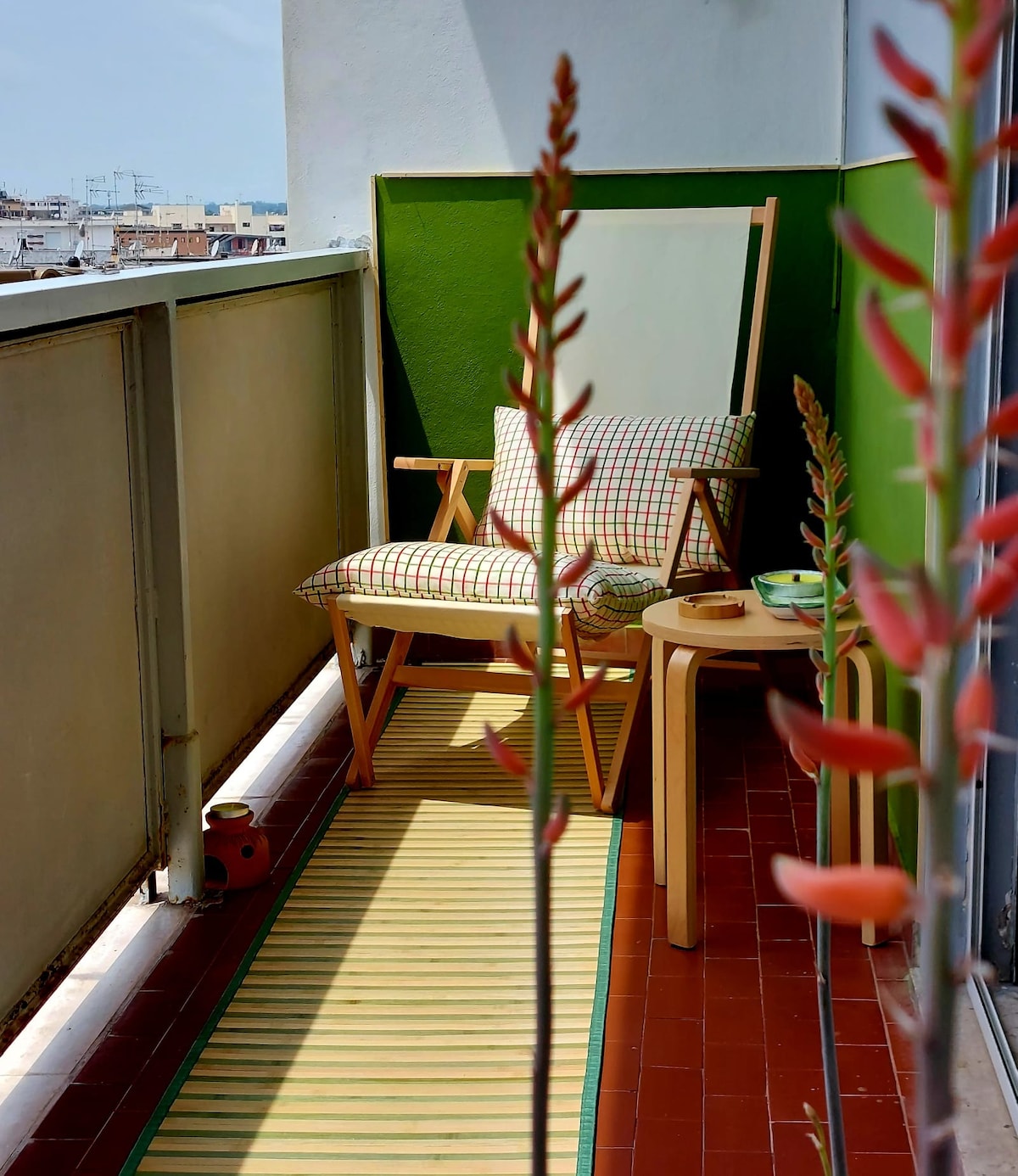 A balcony is shown, featuring a wooden chair with a cushion and a small round table. Bright green walls frame the space, while a striped mat runs along the floor. Natural plants add a touch of color and vibrancy to the setting.