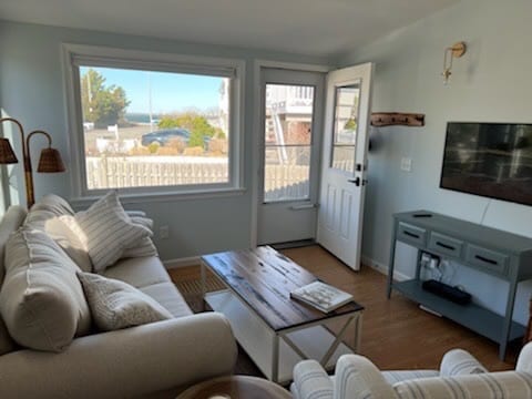 A light-filled living area features a comfortable sectional sofa with neutral-colored cushions, positioned near a large picture window. A wooden coffee table sits in the center, with a small console table in the background housing a television, adding functionality to the inviting space.
