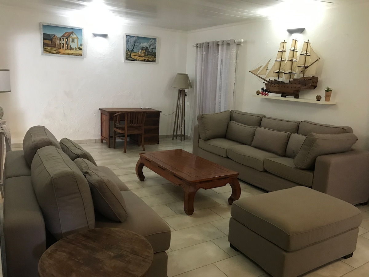 A spacious living area is equipped with light-toned sofas arranged around a wooden coffee table. A wooden desk is positioned nearby, accompanied by a lamp. Wall art depicting rural scenes adds a decorative touch, while a model ship is displayed on a shelf.