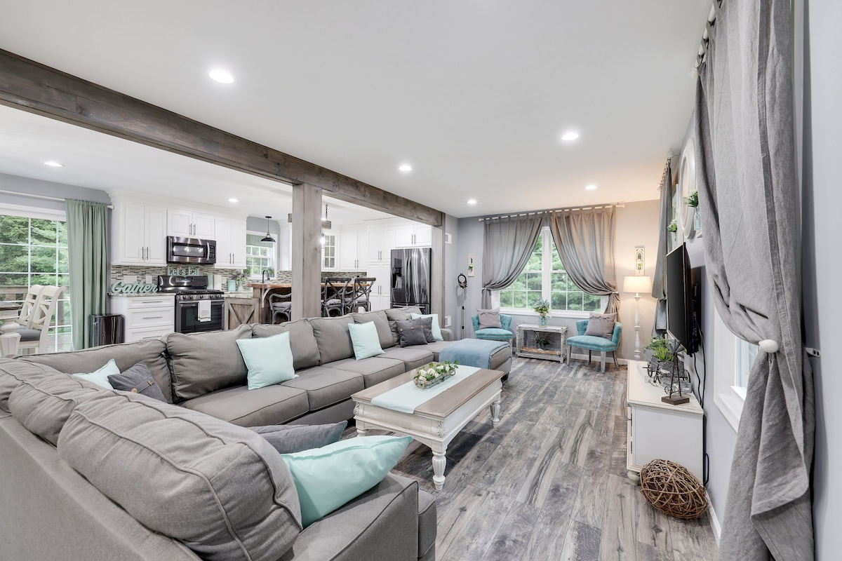 A spacious living area is featured, with a large sectional sofa adorned with light blue cushions. Natural light streams through large windows, illuminating the open concept layout that connects the living area to the kitchen, showcasing modern appliances and a cozy dining space.