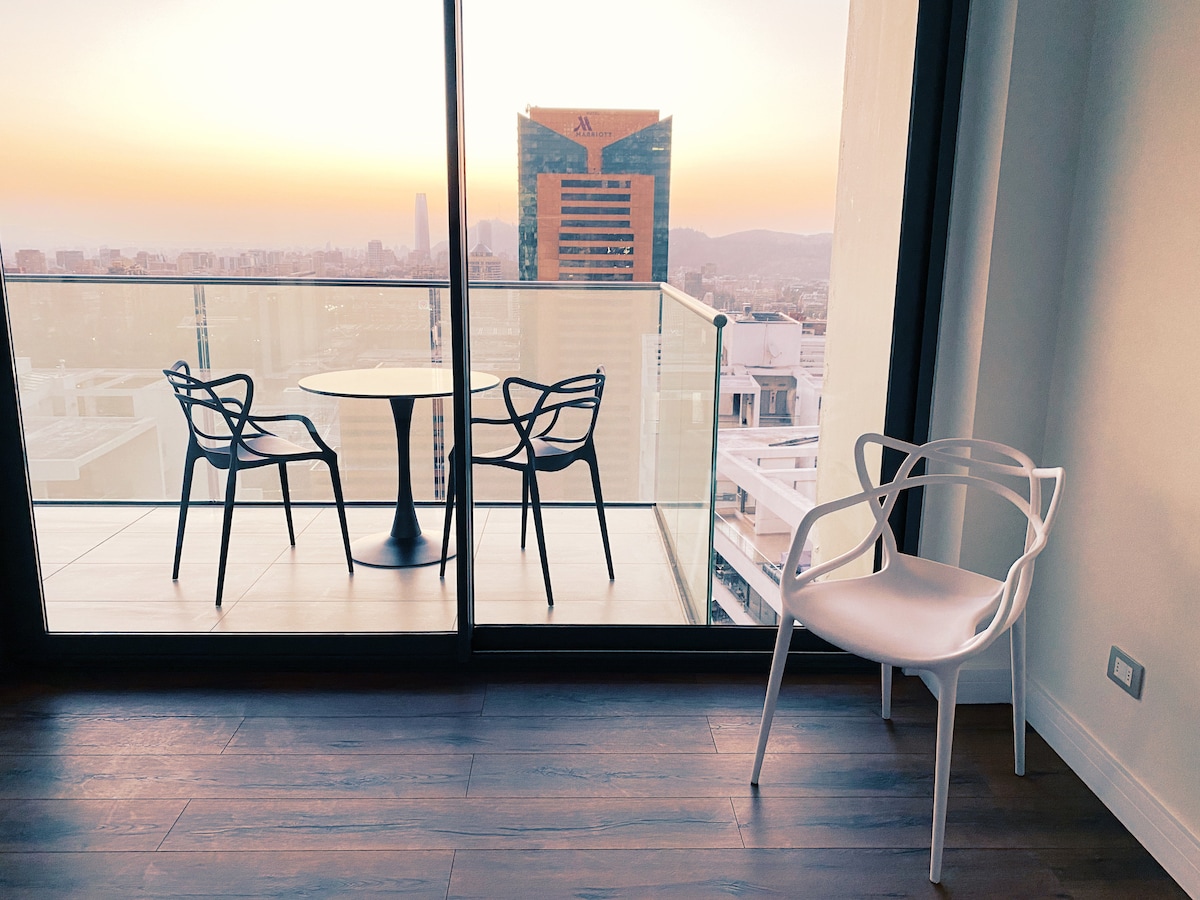 A balcony view showcases modern outdoor furniture, including a round table and two sleek chairs. Floor-to-ceiling glass doors offer a panoramic backdrop of the sunset over the city skyline, enhancing the space's bright and spacious atmosphere.