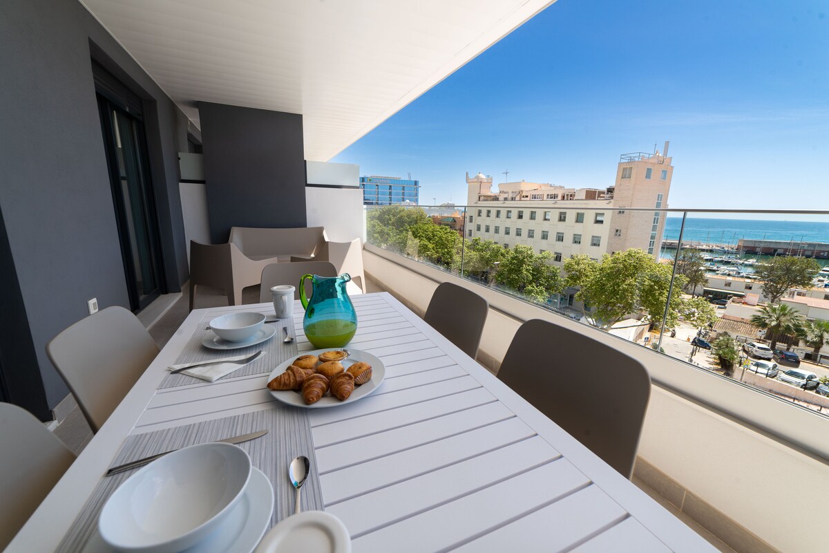 A spacious balcony table is set for breakfast with plates, cutlery, and a vibrant pitcher next to a plate of pastries. The view shows a bustling street and the coastline, framed by clear blue skies and nearby buildings.