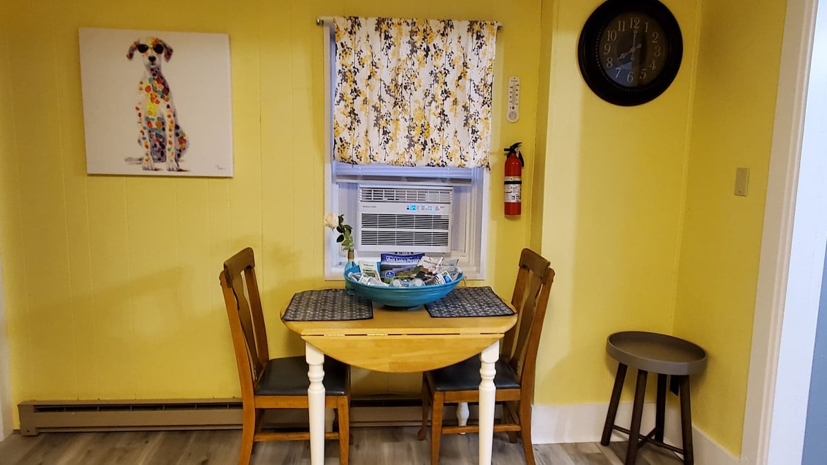 A small dining area is present with a round wooden table and two chairs, complemented by a decorative bowl at the center. A colorful dog painting hangs on the wall above, while a window is adorned with floral curtains. A fire extinguisher is mounted nearby.