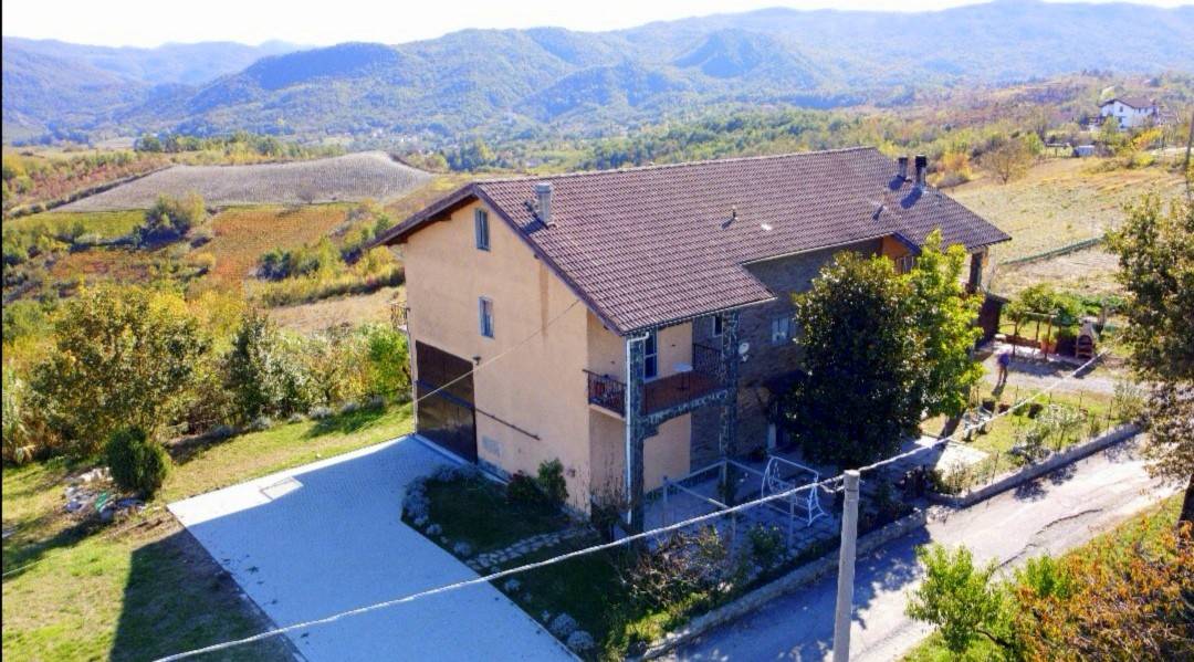 A two-story house is situated in a rural landscape, surrounded by rolling hills. The exterior features a combination of stone and smooth surfaces. A spacious gravel area is visible in front, with greenery and trees surrounding the property, offering a sense of tranquility.