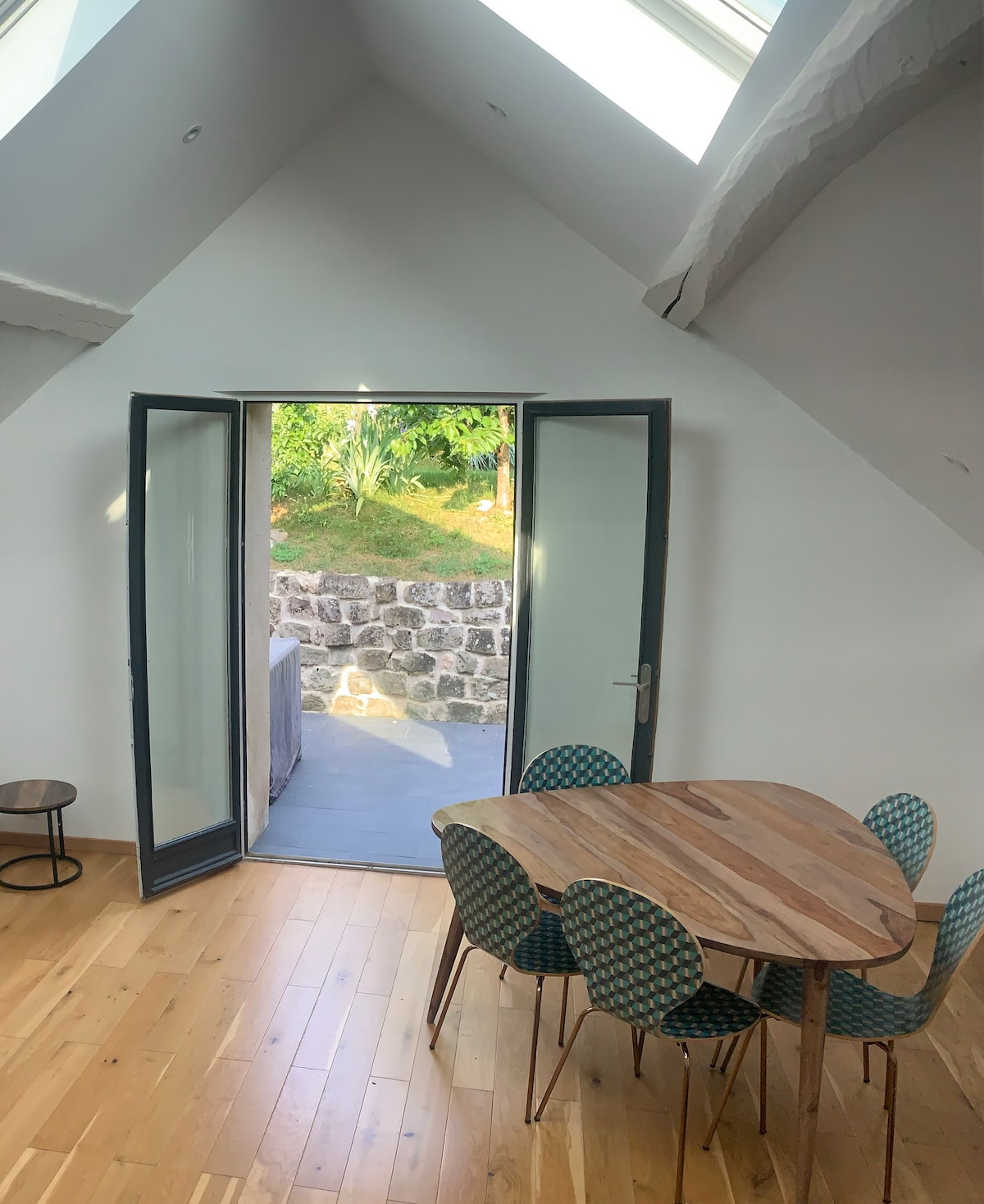 A bright dining area features a wooden table surrounded by patterned green chairs. Double doors open to reveal an outdoor space with greenery, allowing natural light to enhance the room's atmosphere. A small side table is positioned nearby on the light wooden floor.