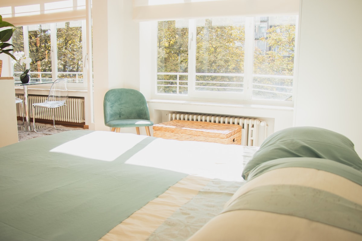 A spacious sleeping area is highlighted by a light-colored bedspread. Natural light floods in through large windows overlooking greenery. A cozy chair and a wicker chest are visible, adding functionality to the bright, open space.