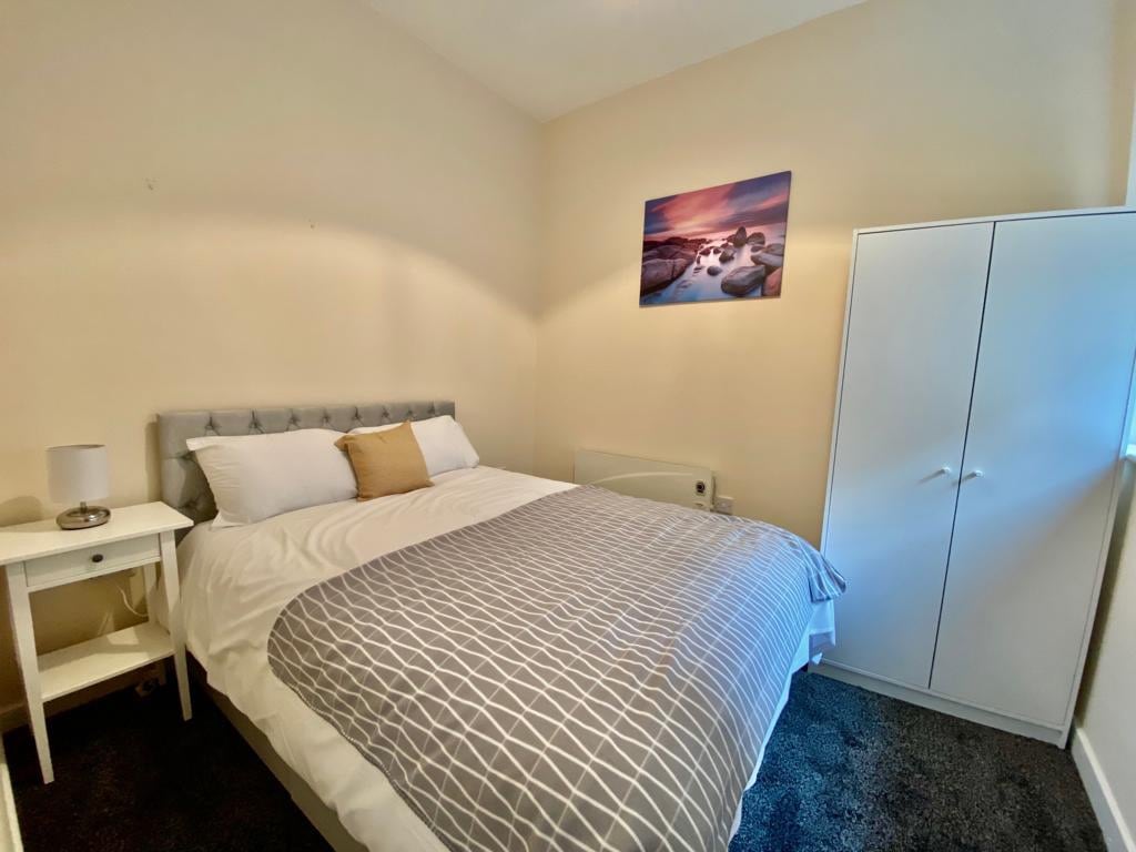 A cozy bedroom is highlighted by a spacious bed dressed in crisp white linens and a grey patterned blanket. A nightstand with a lamp sits beside the bed. A wardrobe is positioned against the wall, while artwork depicting a serene landscape adorns the light-colored wall.