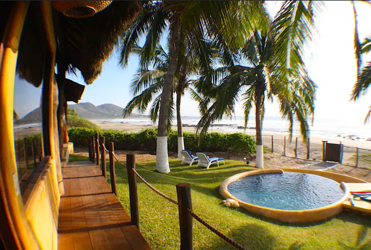 A serene outdoor space features well-maintained grass surrounded by palm trees. A small circular pool is visible, with two lounge chairs placed nearby. The beach can be seen in the background, with gentle waves lapping at the shore.