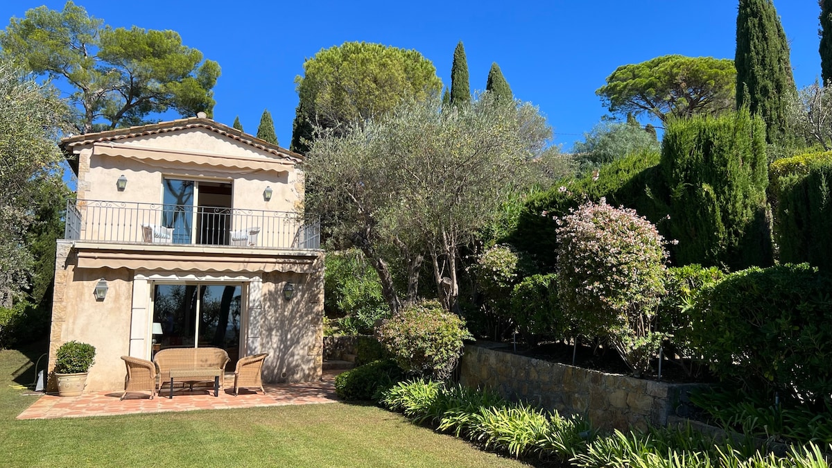 A charming house with a balcony overlooks a well-maintained garden. Various types of greenery are visible, including tall trees and neatly trimmed bushes. The outdoor seating area is positioned on a tiled patio, providing a cozy spot to relax amidst the serene landscape.