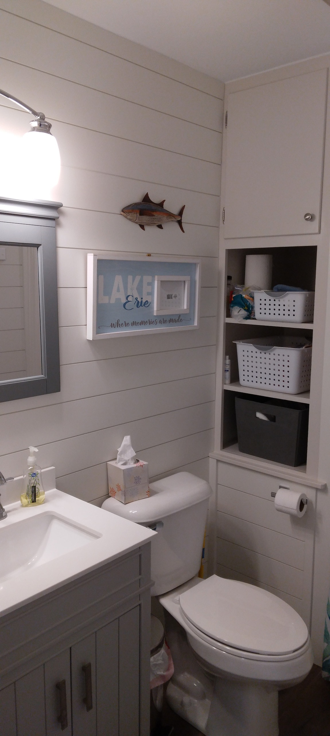 A bathroom features shiplap walls, complemented by a large mirror above a neatly arranged vanity. Shelving units are visible, containing storage baskets. A decorative piece related to the lake is mounted on the wall, adding a subtle thematic touch.