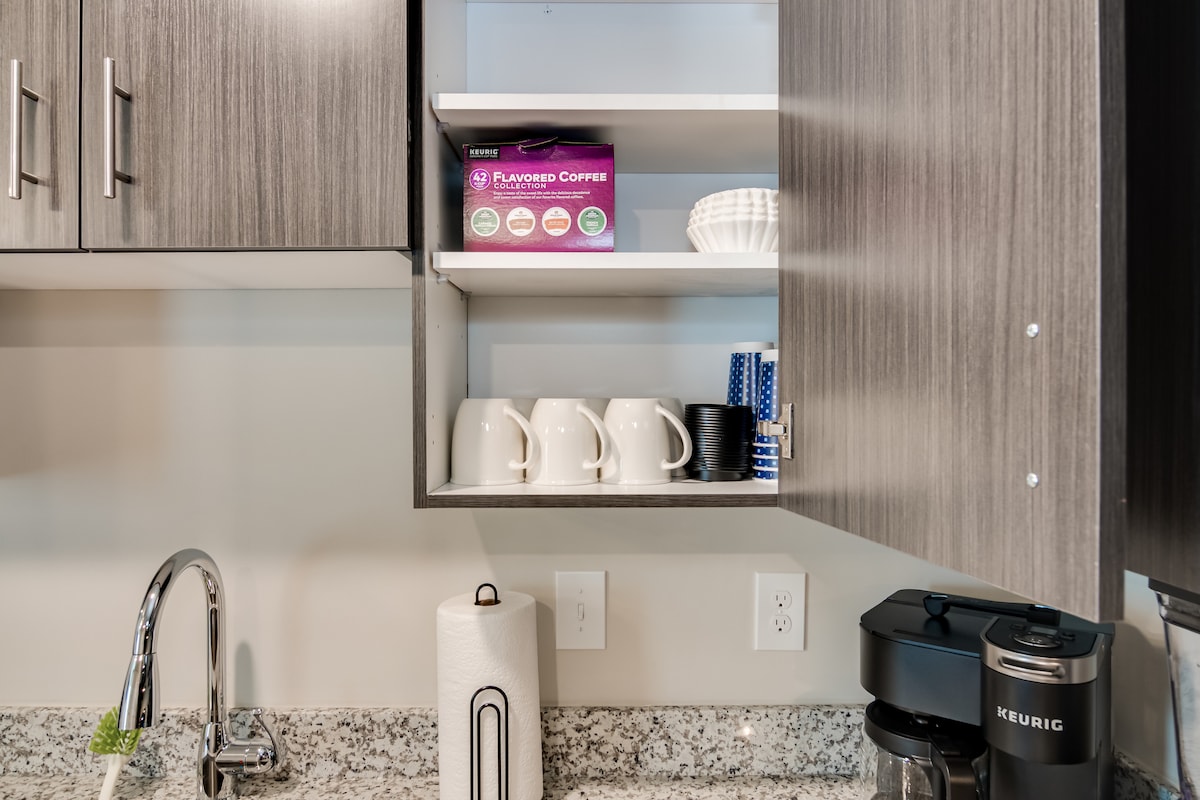 A kitchen cabinet is shown with neatly arranged coffee mugs and a box of flavored coffee pods. Below, a drip coffee machine and a roll of paper towels are visible on the counter, surrounded by a granite surface.