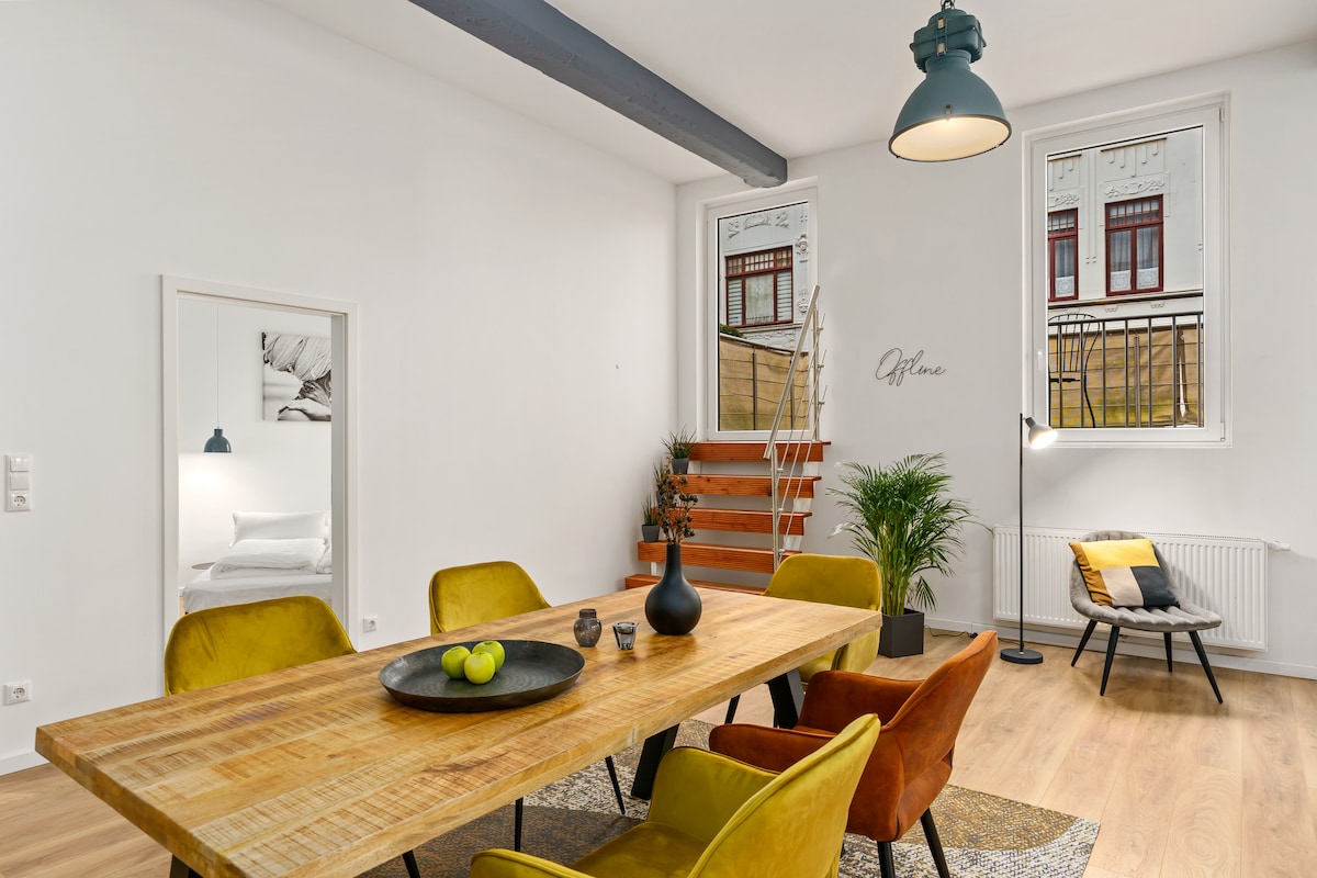 A spacious dining area features a large wooden table surrounded by vibrant yellow chairs. Large windows offer natural light along with a view of the street. A potted plant adds a touch of greenery, and a floor lamp provides additional illumination.
