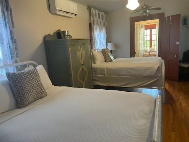 The room contains two full-size beds with white linens. A decorative pillow is placed on one bed. A green cabinet sits against the wall, and windows with light curtains allow natural light to enter. Wooden flooring adds warmth to the space.