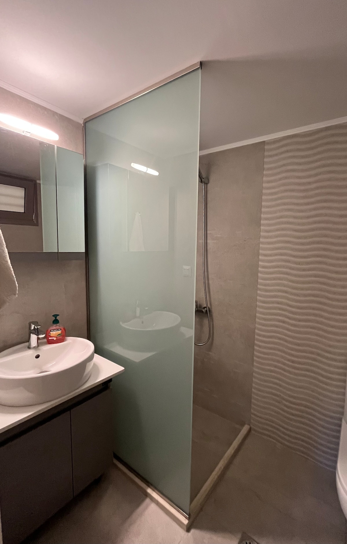 A modern bathroom is featured, showcasing a glass shower enclosure next to a sleek vanity with a round sink. Reflective surfaces and neutral tile tones enhance the space's contemporary feel. A towel hangs on a nearby hook, and natural light illuminates the room.