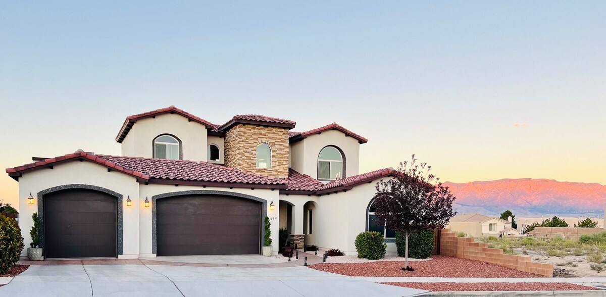 The exterior of a two-story tuscan-style home features a stone and stucco facade, terracotta roof tiles, and arched windows. A paved driveway leads to a three-car garage. Desert landscaping surrounds the entrance, complemented by a backdrop of mountains under a soft twilight sky.