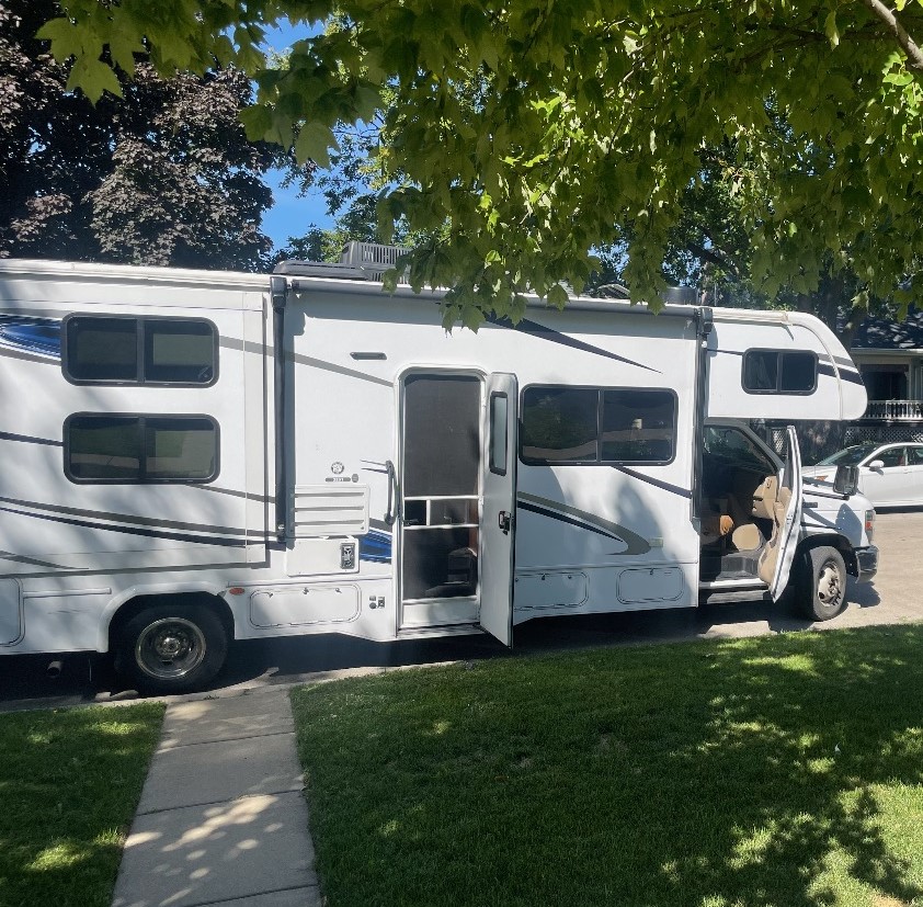 A parked RV is shown, featuring a side door that is open to reveal a welcoming entry. The vehicle is surrounded by green trees and grass, with sunlight casting soft shadows. Multiple windows on the RV provide natural light to the interior.