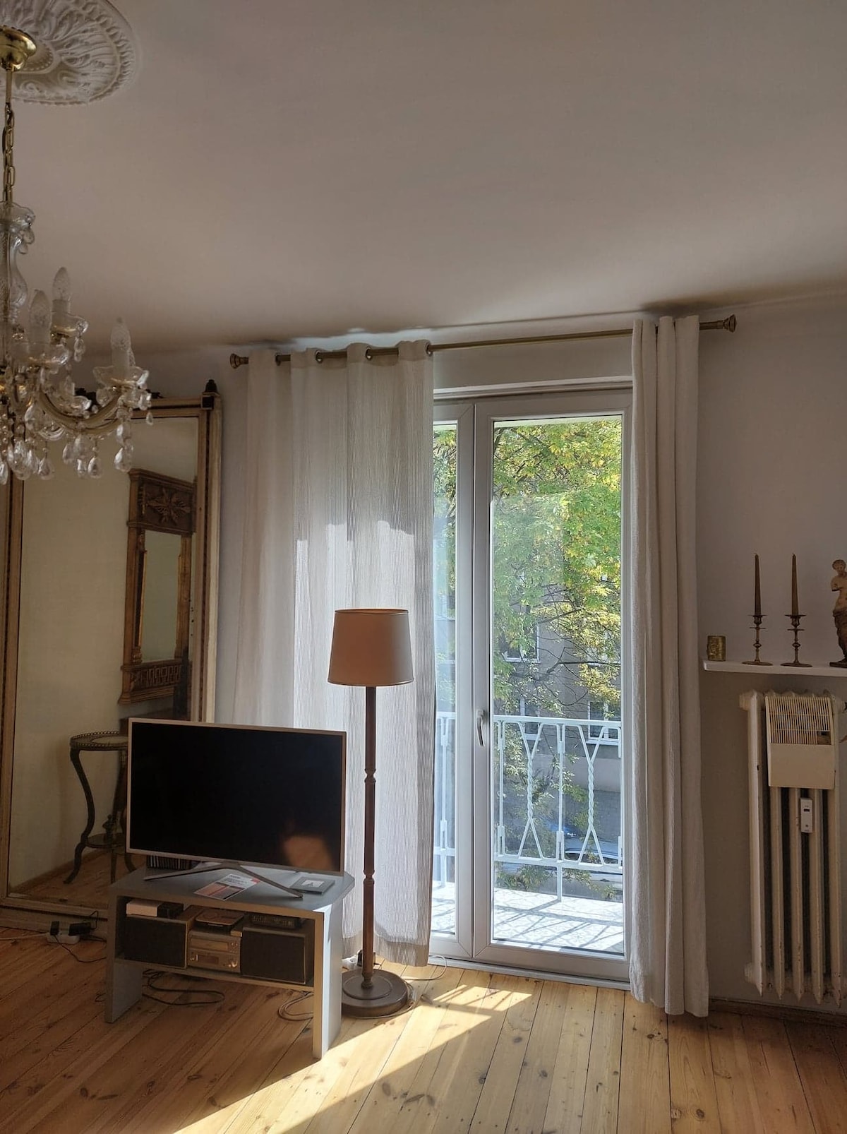 A bright living space is showcased, featuring a large glass door that leads to a balcony. Sheer white curtains frame the door, allowing natural light to filter in. A TV is positioned on a small stand, while stylish decor items are visible throughout the room.