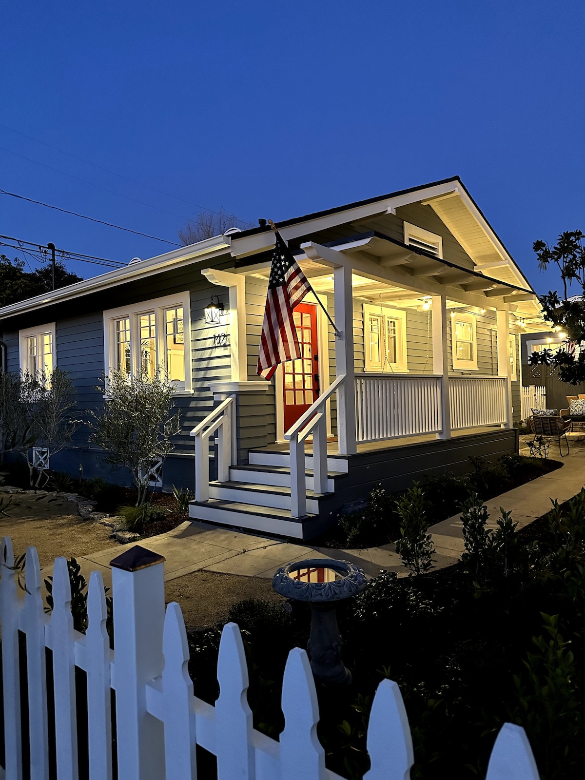 The exterior of a bungalow is shown at dusk, featuring a welcoming front porch with white railing and steps leading up to a vibrant red door. An American flag hangs beside the entrance, and the home's charming lights create a warm glow against the blue sky.