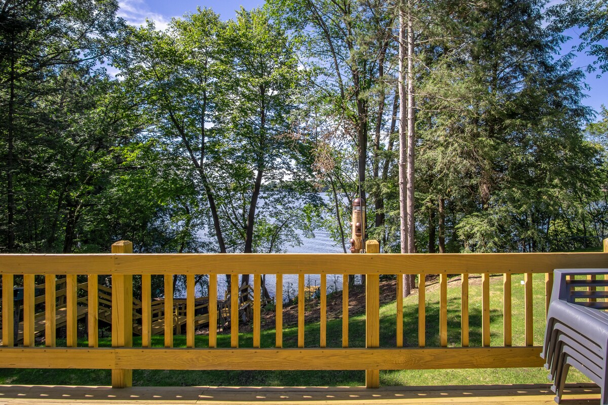 A wooden deck railing captures a view of lush greenery leading down to a serene lake. Sunlight filters through the trees, highlighting the calm water beyond, creating a peaceful natural setting.