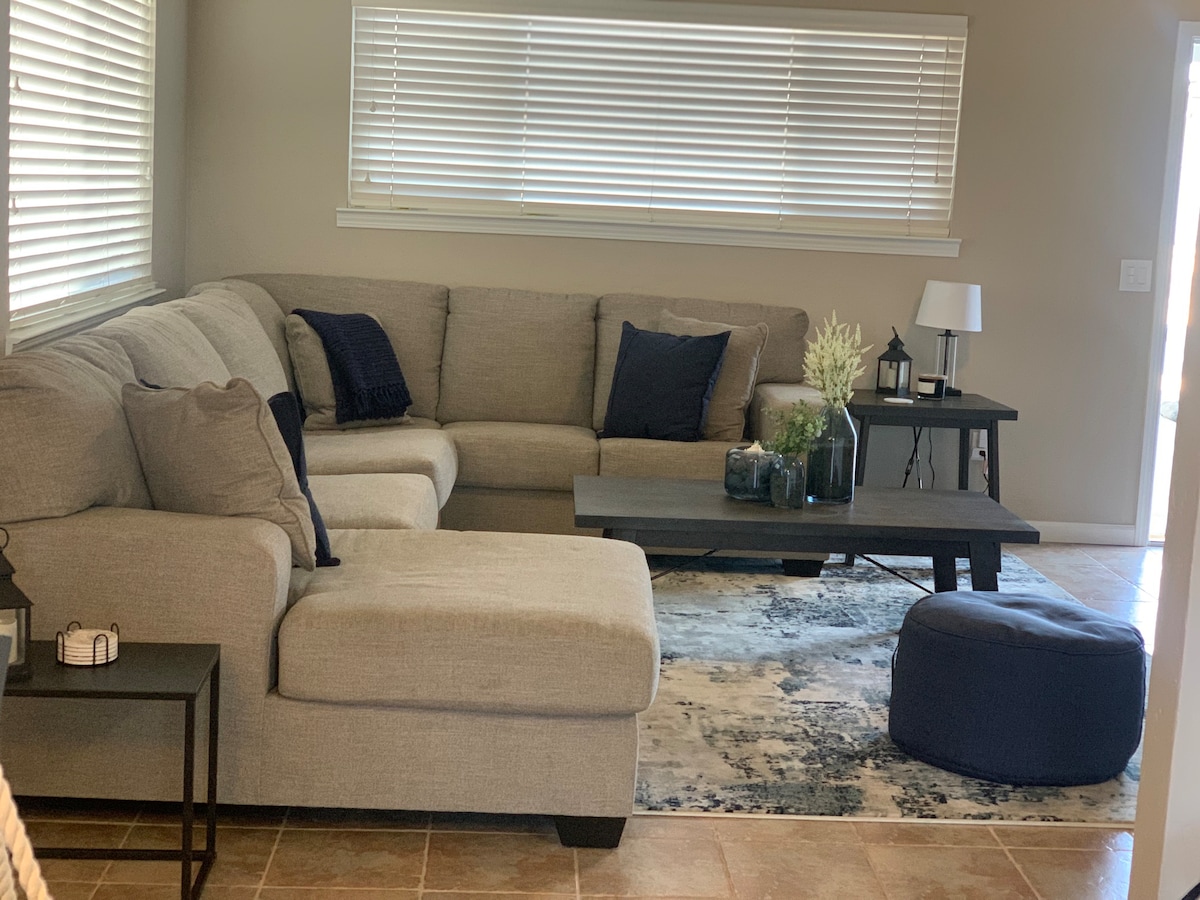 A spacious living area is furnished with a light grey sectional couch adorned with navy blue pillows. A dark coffee table sits atop a patterned area rug, complemented by decorative plants. A small lamp and a round pouf enhance the welcoming ambiance of the room.
