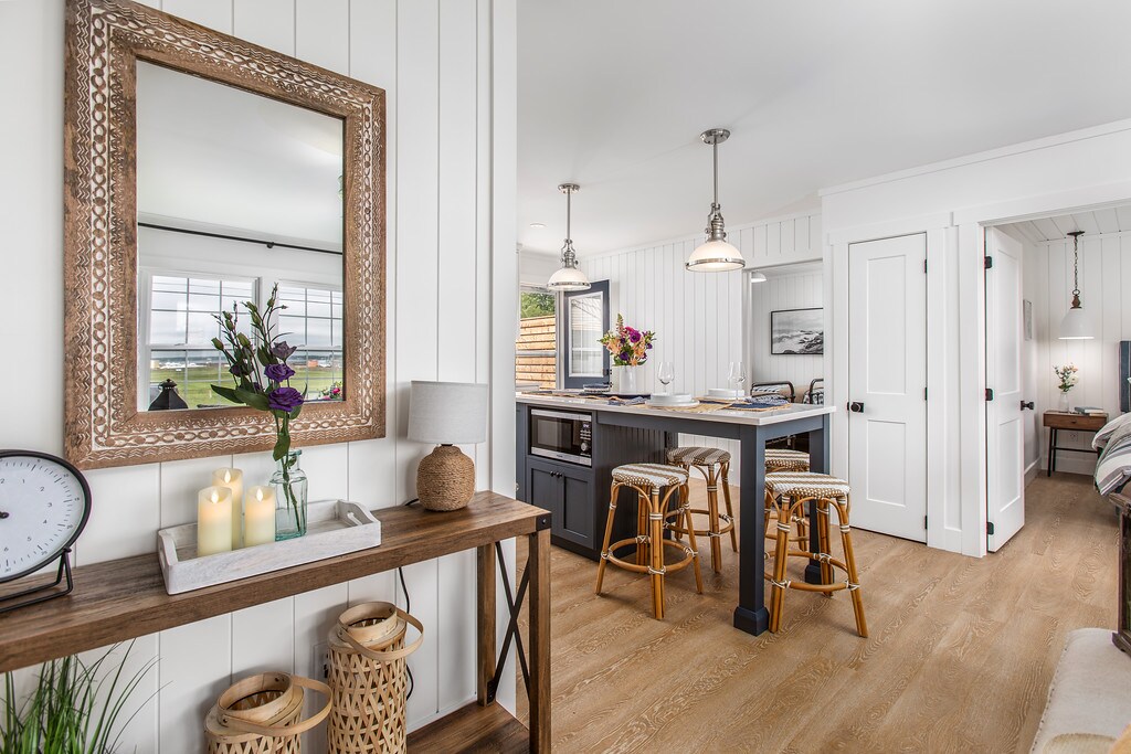 An open kitchen space is depicted, featuring a large island with three wooden bar stools. A mirror with an ornate frame hangs on the wall, reflecting light. Decorative elements include a vase with fresh flowers and a selection of candles, creating a welcoming ambiance.