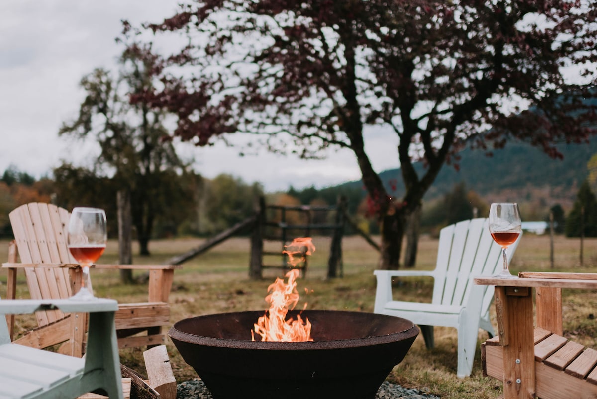A firepit is surrounded by wooden seating in a tranquil outdoor setting. Glasses of rose wine on side tables complement the inviting atmosphere. In the background, a tree and rolling fields create a peaceful backdrop, enhancing the outdoor experience.