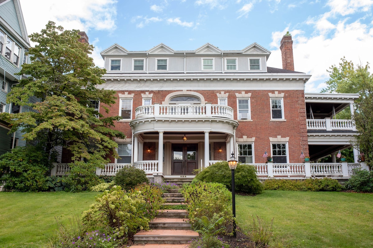 The historic mansion showcases a grand facade with multiple levels, large windows, and a welcoming porch supported by columns. Lush greenery and landscaping line the pathway leading to the entrance, enhancing the elegant and classic architecture.