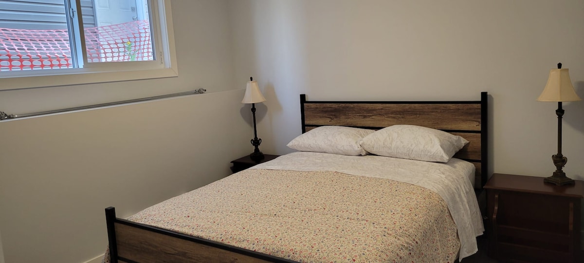 A cozy bedroom features a spacious bed with a light quilt atop it. Two bedside tables with lamps are positioned on either side, while natural light floods through a nearby window.