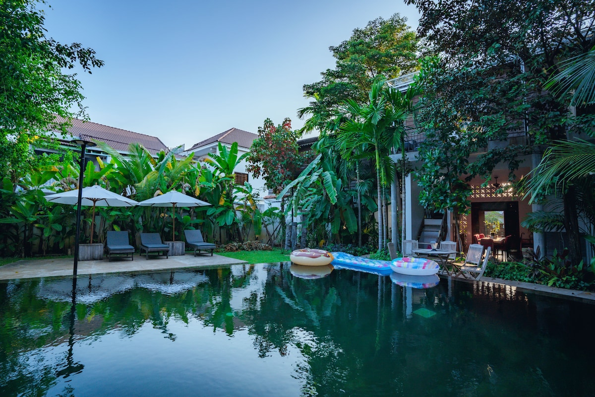 A serene outdoor area highlights a tranquil pool surrounded by lush greenery. Comfortable lounge chairs are positioned under shaded umbrellas. Colorful inflatable floats are placed in the water, inviting relaxation in a peaceful tropical setting.
