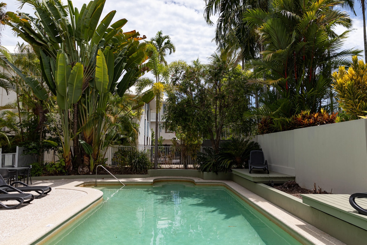 A serene swimming pool is surrounded by lush tropical foliage, creating a tranquil atmosphere. Sun loungers are positioned nearby, offering a place to relax. The clear water reflects the sky, inviting guests to enjoy a refreshing dip.