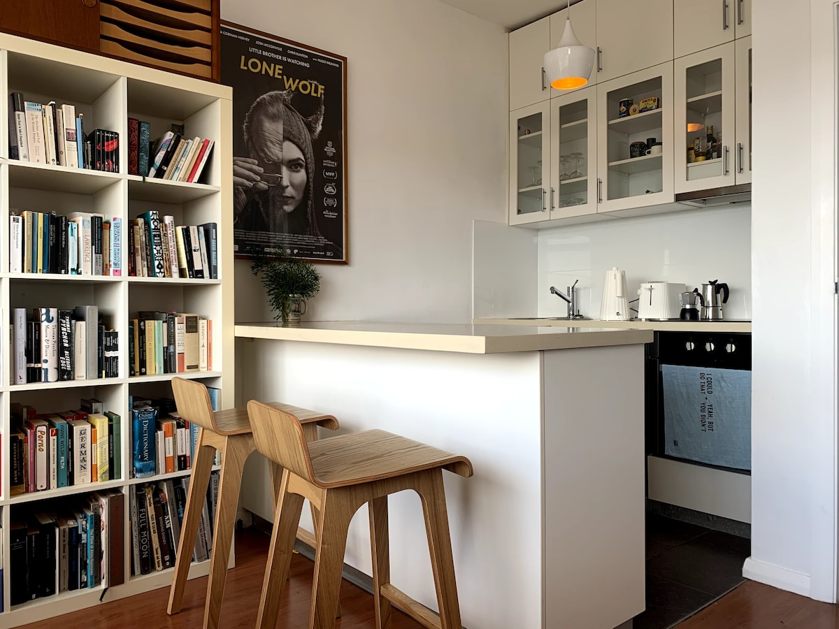 A compact kitchen area features modern appliances, including a dishwasher and coffee maker. A light-colored breakfast bar with two wooden stools provides seating. A tall bookshelf filled with various books stands nearby, and a movie poster is displayed on the wall.