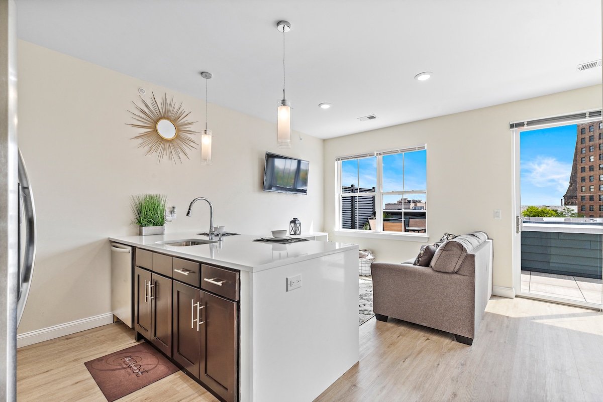 A modern kitchen is visible with sleek cabinetry and a spacious countertop. Natural light floods the area through large windows, providing views of the city skyline. A comfortable seating area and flat-screen TV are included, enhancing the contemporary design of the space.