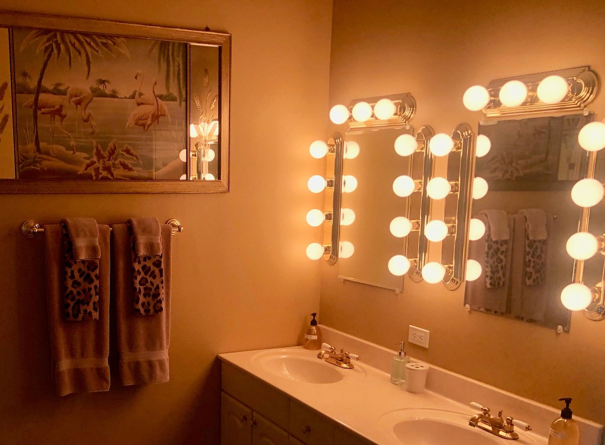 A well-lit bathroom features two mirrors surrounded by round bulbs, enhancing visibility. Soft beige walls are complemented by a framed artwork depicting flamingos. Two plush towels hang on a bar, and decorative soap and toiletries are neatly arranged on the counter.