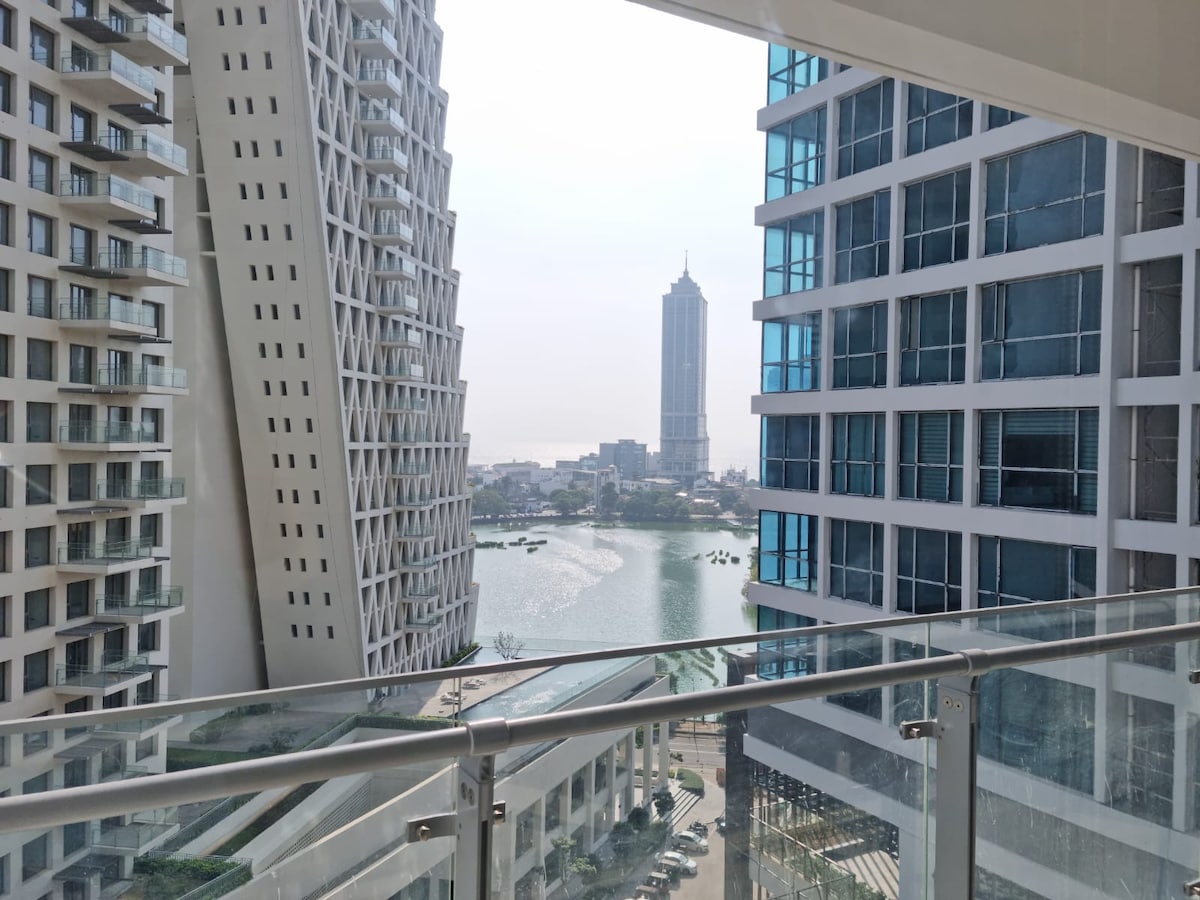 A modern balcony view showcases surrounding high-rise buildings and a serene body of water, with city landmarks visible in the background. The clear glass railing offers unobstructed sightlines, allowing for ample natural light and an expansive view of the urban landscape.