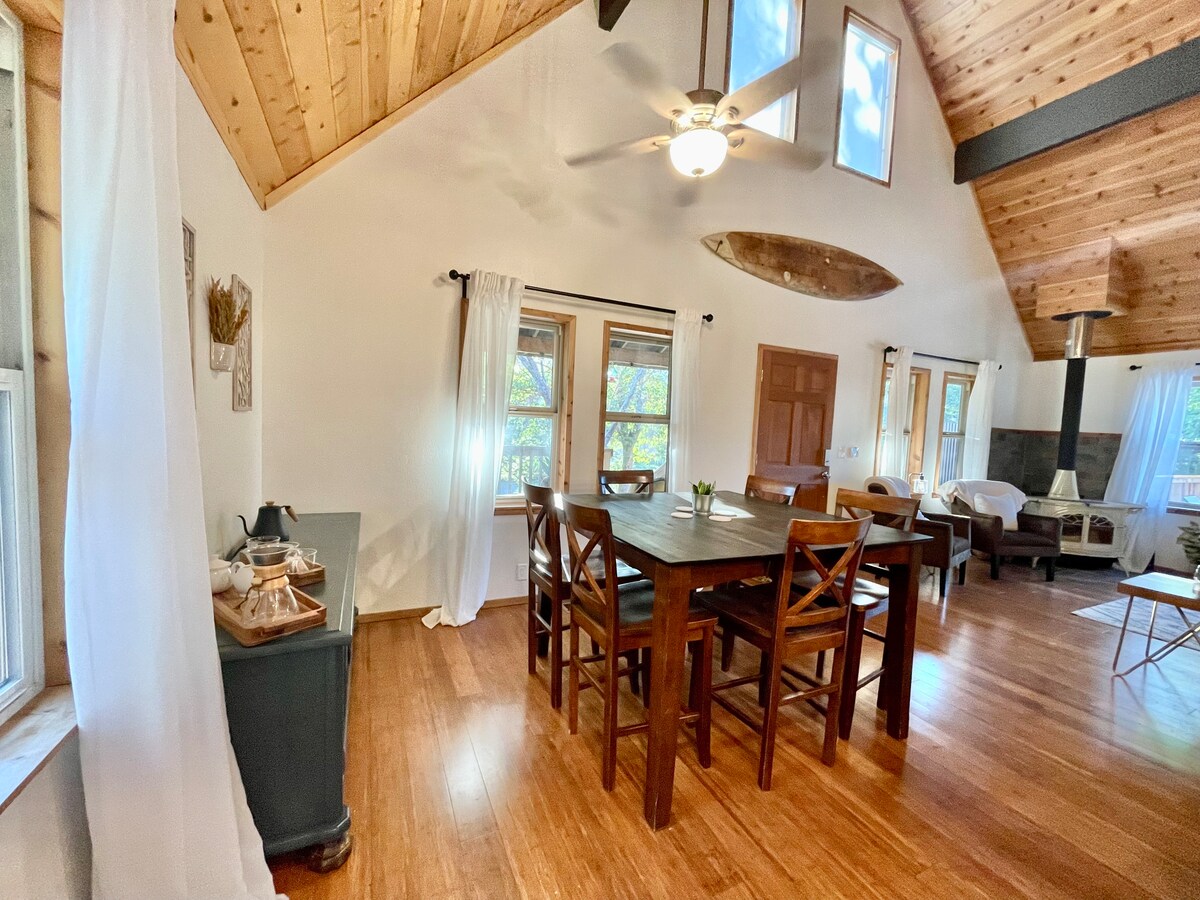 An open-concept living space features high vaulted ceilings with wooden beams and large windows allowing natural light to fill the room. A wooden dining table is surrounded by chairs, with a cozy seating area visible in the background, showcasing a vintage wood stove.